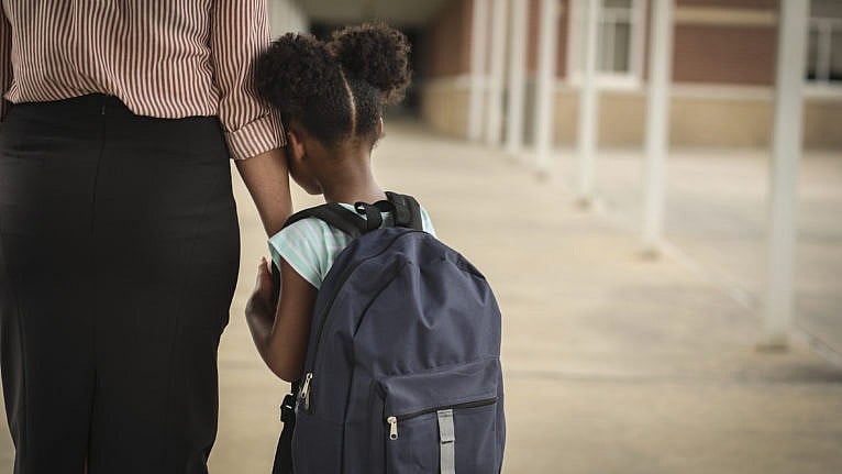 Elementary age, African American girl holds mom or teacher's hand before school begins. She wears a backpack and clings to mom with uncertainty about starting school.