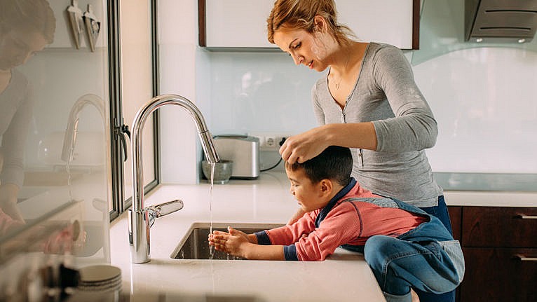 A kid washes their hands at the kitchen sink while their mom watches.
