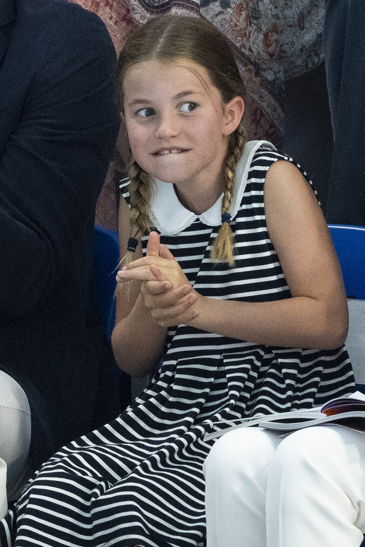 Princess Charlotte makes a funny face while sitting in the stands at the Commonwealth Games wearing a striped collared dress and braided pigtails.