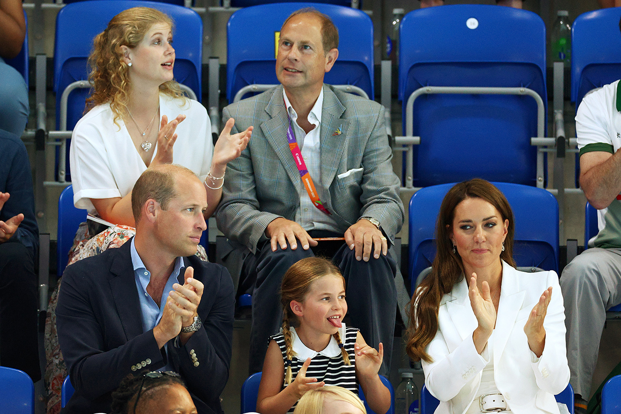 Princess Charlotte sticks out her tongue while sitting in the stands between her parents Prince William and Kate MIddleton at a Commonwealth Games swim meet.