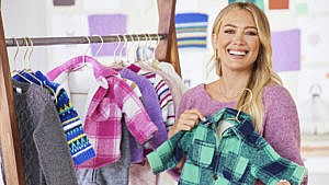 Hilary Duff holding up a baby-sized green plaid shacket with a rack of baby clothes behind her.
