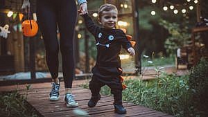 Toddler wearing black and orange costume walks with adult holding hand and trick-or-treating basket on a dark evening