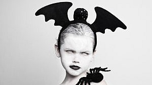 A black and white photo of a little girl without iris wearing a bat head band and a toy spider on her shoulder.