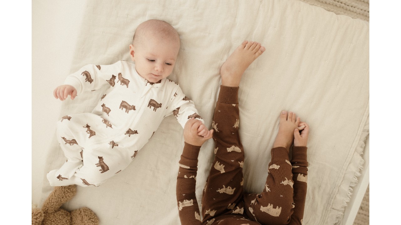 a baby and toddler rest on a beige blanket, wearing white and rust pyjamas with bear cub print