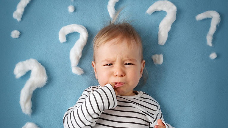 A baby with a finger in their mouth has a disgruntled look on their face. around their head are question marks made of cotton balls