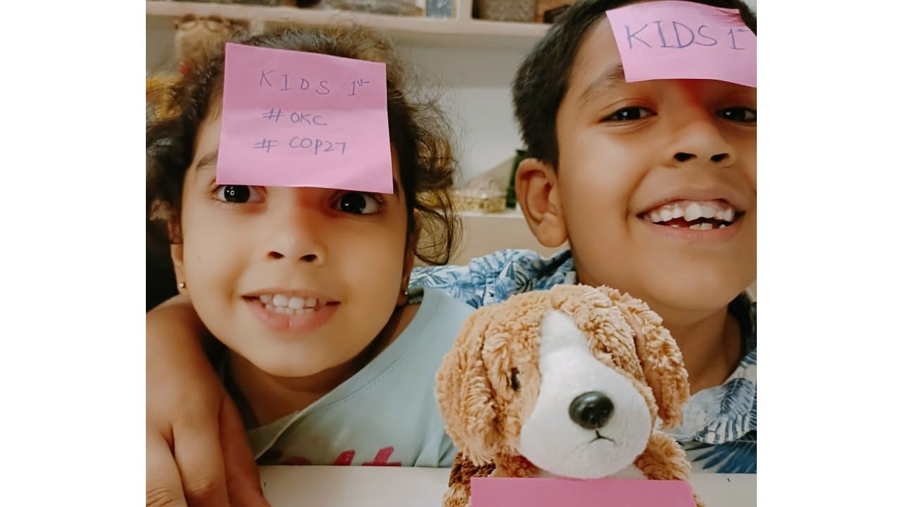 Two young children have pink post it notes on their foreheads, reading #KidsFirst #COP27, and their stuffed puppy does too