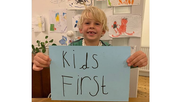 Young child holds up a hand-written sign reading Kids First in front of a family room with kid drawn pictures on the wall