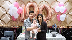 A picture of a family holding their baby in front of balloons that forms the number 100.