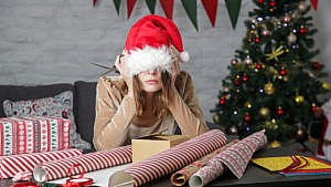 A women with a santa hat covering her eyes is frustrated as she is surrounded by christmas gift wrappers.